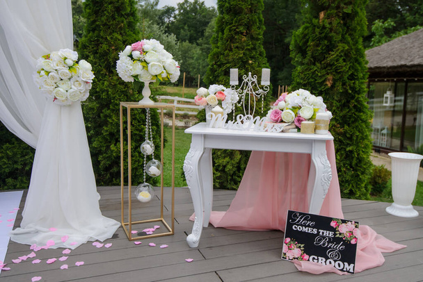 Een witte vintage houten tafel bij een trouwboog met boeketten van bloemen, kaarsen en roze gordijnen met op een groene achtergrond - Foto, afbeelding