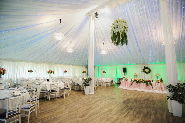 El interior de una sala de banquetes de boda: mesas redondas con mantel blanco y todos los suministros necesarios para la cena en ellos, ramo de flores; cortinas blancas, piso beige, sillas blancas, mesa presidium con mantel rosa
 - Foto, imagen