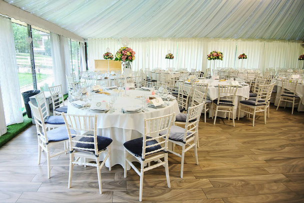 Interiér svatebního hostinského sálu: kulaté stoly s bílým ubrusem a vším potřebným k večeři, kytice květin; bílé záclony, béžová podlaha, bílé židle s modrými polštáři - Fotografie, Obrázek