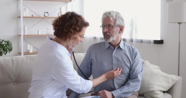 Female doctor listening old patient with stethoscope during homecare visit - Felvétel, videó