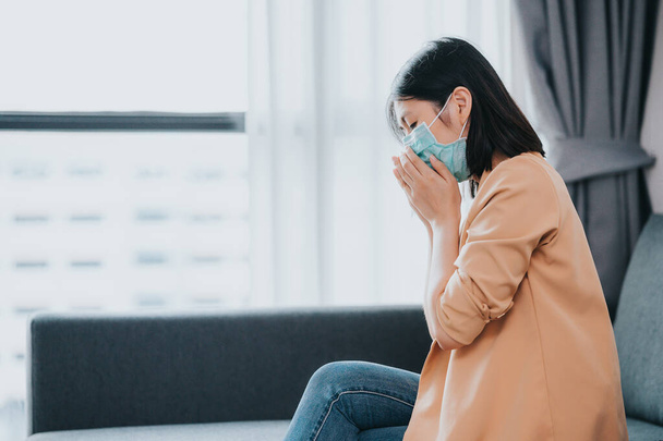 Asian woman wearing face mask to protect from coronavirus flu virus and disease coughing and feeling sick in living room - Фото, изображение