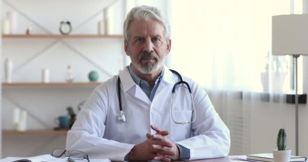 Reliable smiling older professional physician sitting at workplace, portrait - Metraje, vídeo
