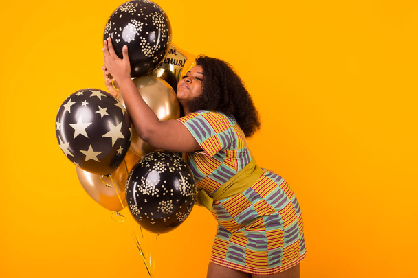 Vacances, fête et concept amusant - Portrait de jeune femme afro-américaine souriante regardant doux sur fond jaune tenant des ballons
. - Photo, image