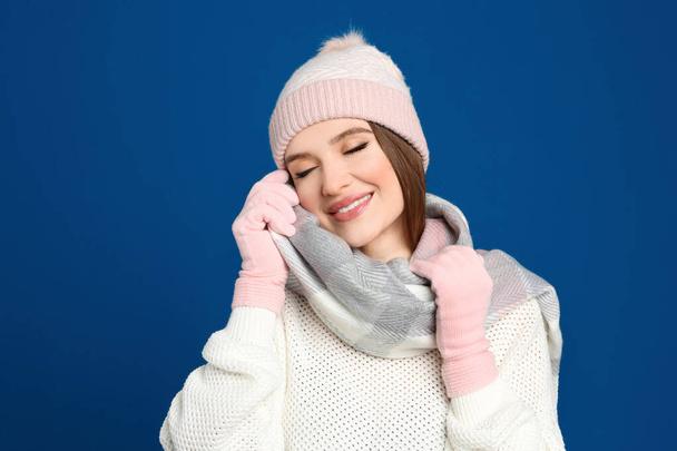 Νεαρή γυναίκα που φοράει ζεστό πουλόβερ, γάντια, κασκόλ και καπέλο σε μπλε φόντο. Χειμερινή περίοδος - Φωτογραφία, εικόνα