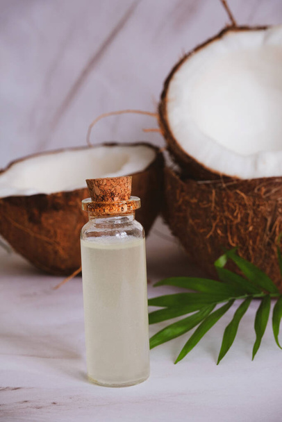 close up selective focus glass bottle with coconut oil on a blurred background of cracked coconuts and palm leaves - Foto, Bild