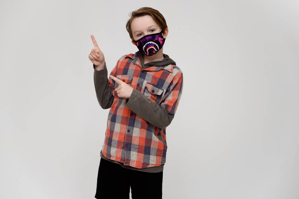 Photo of a masked boy in a plaid shirt on a white background shows his hands to the sides - Фото, изображение