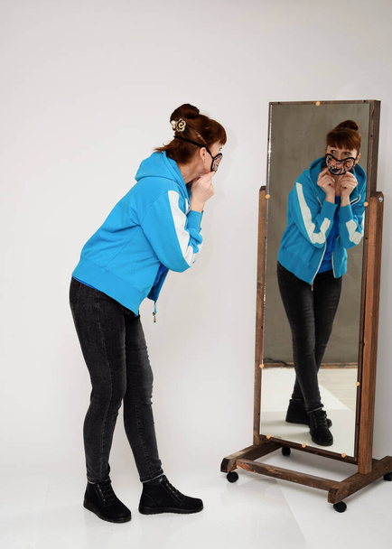 Photo woman in a mask in a blue blouse and jeans on a white background in full growth looks in the mirror - Photo, image
