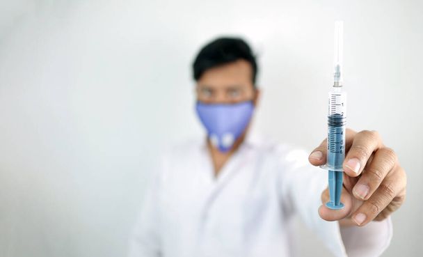 zdravotnický pracovník v bílém plášti a ochranné masce držící injekční stříkačku v ruce se selektivním ostřením v ruce s rozmazaným pozadím - Fotografie, Obrázek