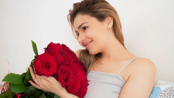 Beautiful woman with red rose flower bouqet. Sensual and attractive female smiling and enjoying flowers at home in bed. Romantic and feminine concept. Handsome portrait shot. - Fotó, kép