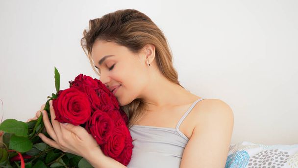 Hermosa mujer con ramo de flores de rosa roja. Sensual y atractiva hembra sonriendo y disfrutando de flores en casa en la cama. Concepto romántico y femenino. Retrato guapo tiro
. - Foto, Imagen