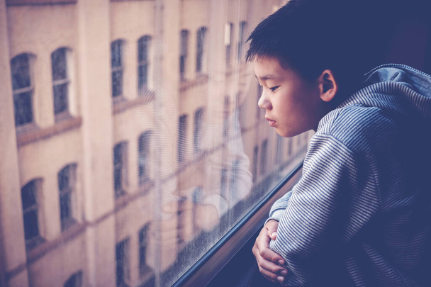 Sad Asian preteen boy feeling lonely, looking out of window from his room, social distancing, isolation, mental health concept - Fotó, kép