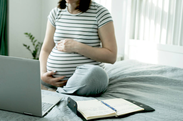 Gyönyörű terhes fiatal nő, aki egy laptopon dolgozik, és az ágyon ül a hálószobában. Otthoni iroda, otthoni oktatás. Társadalmi távolság a karantén alatt, az önelkülönítés fogalma. - Fotó, kép