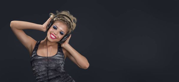 Young beautiful woman portrait listening modern music in headphone isolated over dark gray wide background - 写真・画像