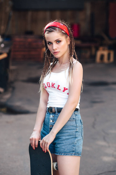 stylish sexy model girl in summer bright casual hipster clothes and red band with skateboard in city - Fotó, kép