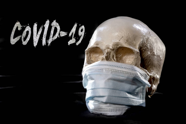 Un cráneo humano con una máscara médica sobre un fondo de textura oscura. Etiqueta: covid-19. Concepto: mortalidad por coronavirus, infección por un virus mortal
. - Foto, Imagen