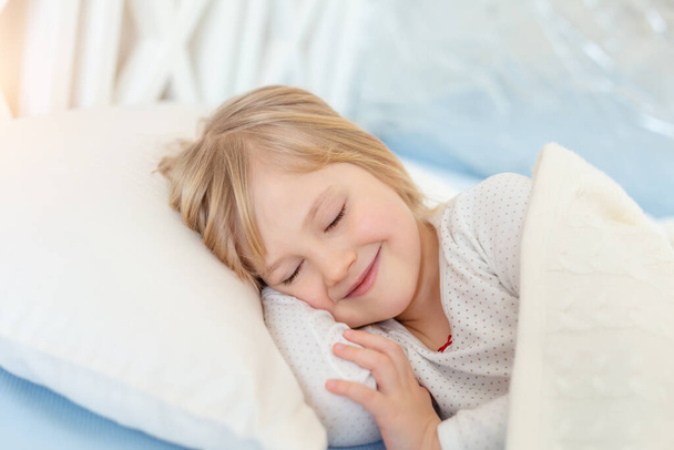 Cute adorable caucasian blond little toddler girl lying in bed on white pillow. Portrait of small beautiful long haired happy baby sweet dreaming asleep and smiling at bedtime in room at home. - Photo, Image