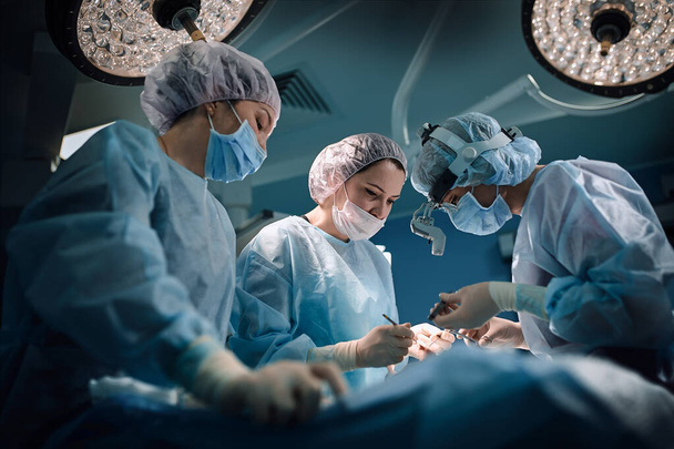 Ιατρική ομάδα στο χειρουργείο, σκοτεινό φόντο. Το θέατρο του χειρουργείου, μια διεθνής ομάδα επαγγελματιών ιατρών σε ένα σύγχρονο χειρουργείο διεξάγουν εγχείρηση. Αποθήκευση - Φωτογραφία, εικόνα