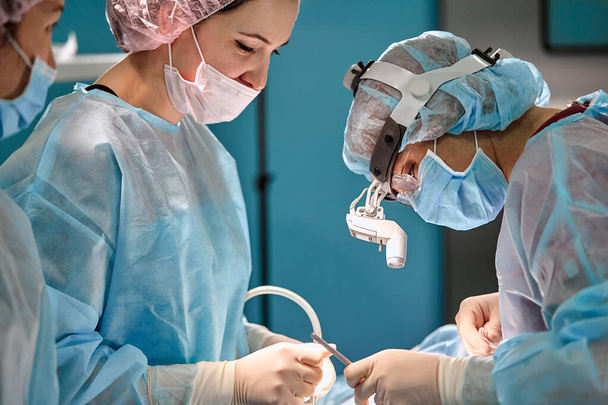 Kirurginen ryhmä leikkaussaliin, lähikuva. Nykyaikaisessa leikkaussalissa työskentelevien lääkäreiden kansainvälinen ryhmä on leikkauksessa. Ihmishenkien pelastaminen, moderni lääketiede, sininen valo
 - Valokuva, kuva