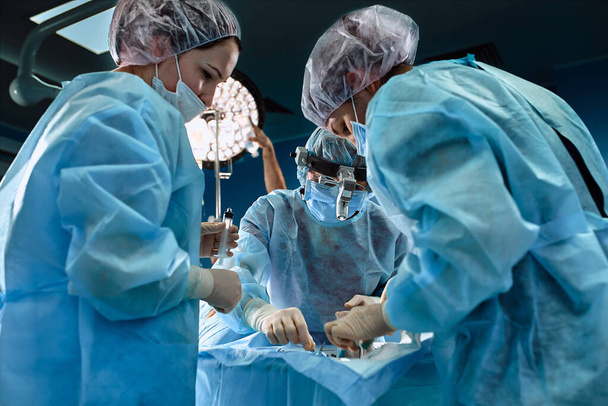 Zespół chirurgiczny na sali operacyjnej, zbliżenie. Międzynarodowy zespół profesjonalnych lekarzy na nowoczesnej sali operacyjnej przechodzi operację. Ratowanie życia, nowoczesna medycyna, niebieskie światło - Zdjęcie, obraz