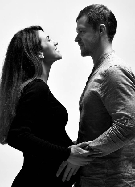 Pregnant girl in black dress and her husband. He hugging her belly, they going to kiss, posing isolated on white. Close up - Fotografie, Obrázek