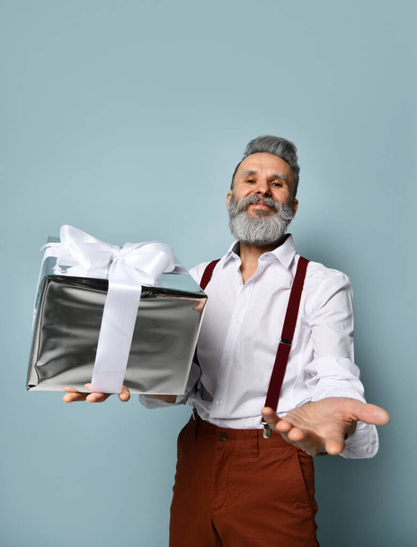 Bearded elderly man in white shirt, brown pants and suspenders. He is smiling, holding silver gift box, posing on blue background - Photo, Image