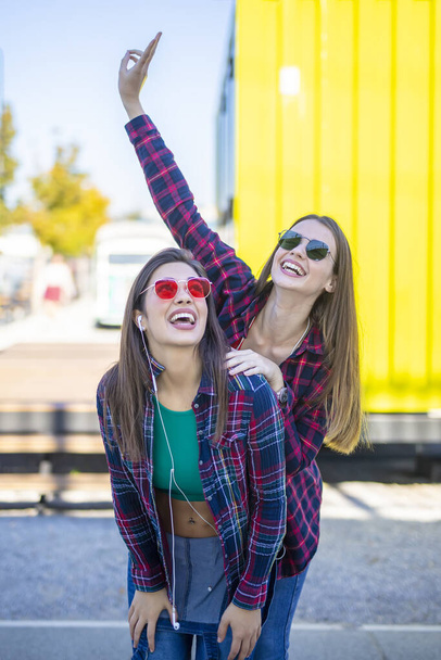 Jovens mulheres com rosto feliz, ao ar livre na rua, ouvir música em fones de ouvido e se divertir
 - Foto, Imagem