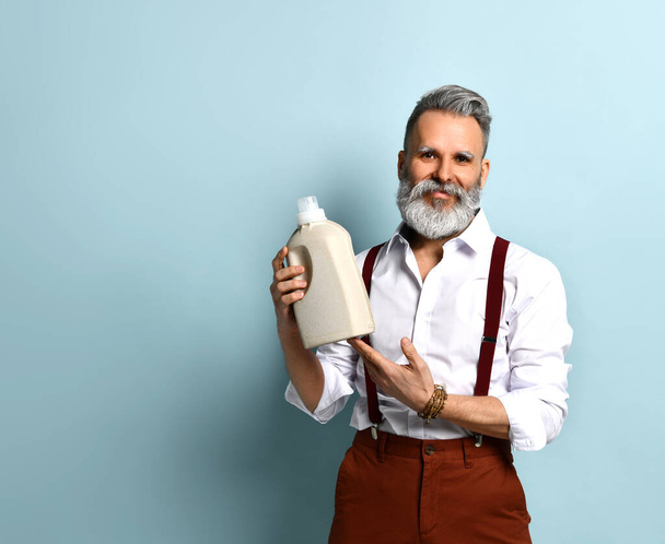 Gray-haired man in white shirt, brown pants. Showing thumb up and bottle mock up of effective detergent, posing on blue background - Valokuva, kuva