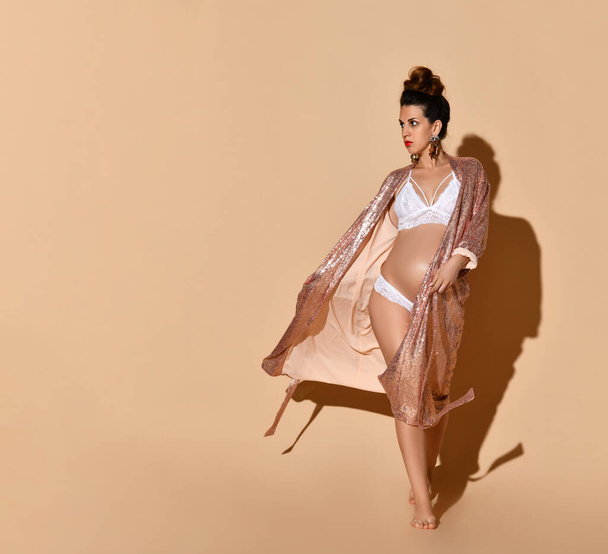 Pregnant female in earrings, white sexy lingerie and golden robe. She is walking against beige background. Full-length, copy space - Φωτογραφία, εικόνα