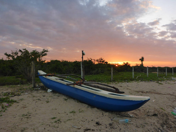 Barco de pesca solitário na areia. tira de moitas tropicais. Acima das moitas, um céu com uma faixa vermelha de pôr-do-sol coberta de nuvens. Tiro no Sri Lanka na costa leste
. - Foto, Imagem
