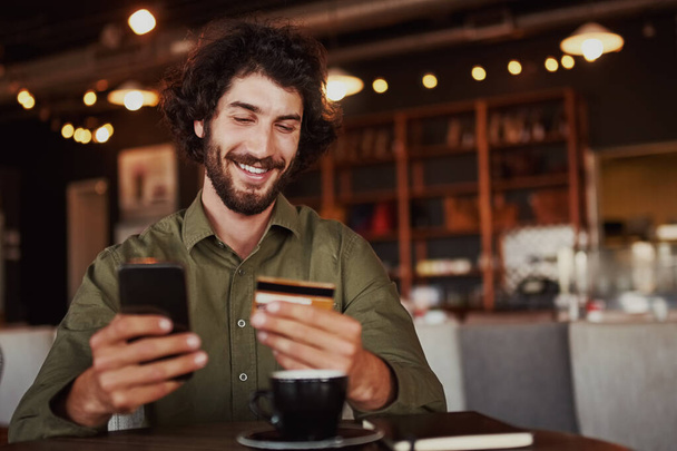 Happy caucasian bearded man holding mobile phone typing card data to make online payment sitting in cafe - Foto, Imagen
