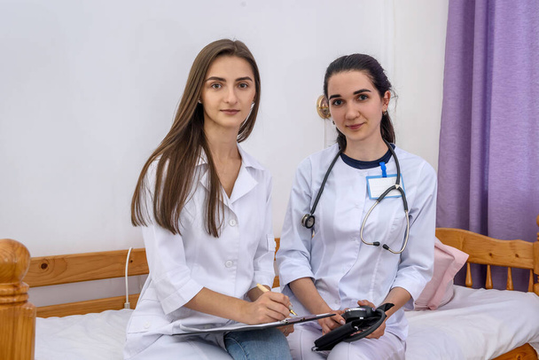 Mujeres jóvenes pasantes sentadas en el hospital en la cama y escribiendo en el portapapeles. Llevan uniformes médicos blancos y un tonómetro.
 - Foto, Imagen