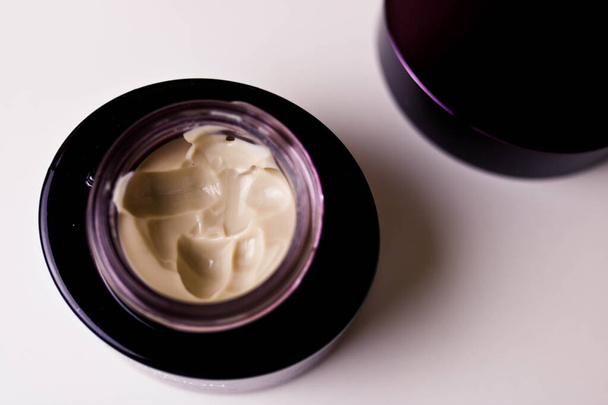 Hidratante crema cosmética remolino en maceta de vidrio púrpura, en la mesa de madera blanca directamente encima. Crema facial en un frasco
. - Foto, Imagen