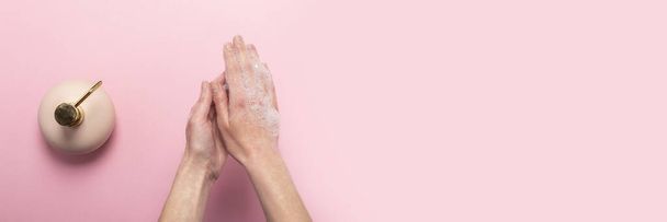 手洗いだ。ピンクの背景に液体石鹸ディスペンサー付きの石鹸女性の手とボトル。手洗いと手の扱いのルールの概念。トップビュー、フラットレイアウト。バナー. - 写真・画像
