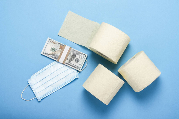 toilet paper rolls, medical mask and dollars banknotes on a blue background. Flat lay, top view. Concept hygiene product, virus, deficiency, quarantine. - Photo, Image