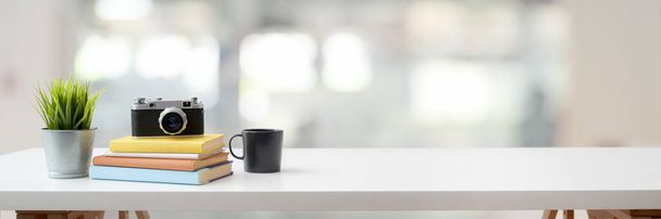 Ausschnittaufnahme eines komfortablen Arbeitsplatzes mit Kamera, Büchern, Kaffeetasse, Baumkännchen und Kopierraum auf weißem Tisch mit unscharfem Hintergrund - Foto, Bild