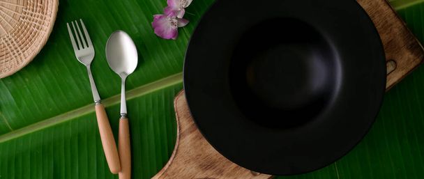 Вид сверху на керамическую тарелку на деревянном подносе с ложкой и фольклор на фоне бананового листа, экологически чистая ресторанная концепция
 - Фото, изображение