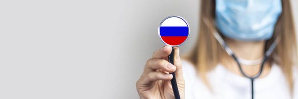 female doctor in a medical mask holds a stethoscope on a light background. Added flag of Russia. Concept medicine, level of medicine, virus, epidemic. Baner. - Photo, Image