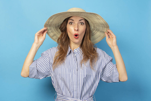joven sorprendida en un vestido y sombrero, sostiene un sombrero con las manos sobre un fondo azul. Concepto de vacaciones, verano y viajes
. - Foto, imagen