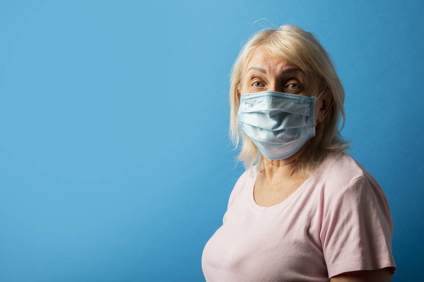 Old woman in protective medical mask on a blue isolated background. The concept of coronavirus, pandemic, no virus, stop, individual protection. Banner. copy space. - Φωτογραφία, εικόνα