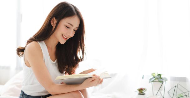 Portrait of smiling happy cheerful beautiful pretty asian woman relaxing reading a book enjoy of rest.Young student girl studying with book in holiday morning vacation on bed at home with copy space - Photo, image