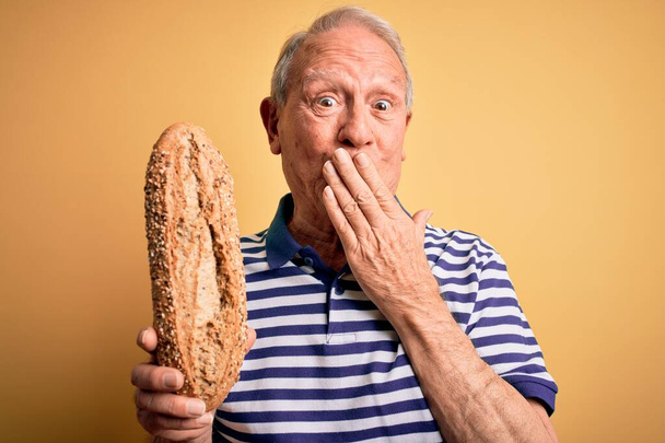 Grey haired senior man holding healthy wholemeal bread over yellow isolated background cover mouth with hand shocked with shame for mistake, expression of fear, scared in silence, secret concept - Photo, Image