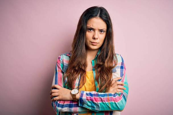 Young beautiful brunette woman wearing casual colorful shirt standing over pink background skeptic and nervous, disapproving expression on face with crossed arms. Negative person. - Photo, Image