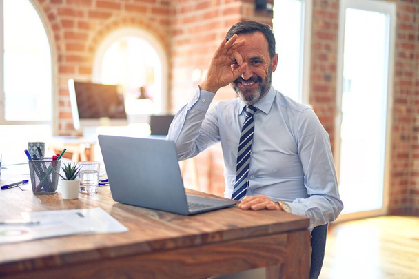 Красивый бизнесмен средних лет в галстуке сидит в офисе, используя ноутбук, делает хорошо жест с улыбкой на руке, глаза смотрят сквозь пальцы с счастливым лицом
. - Фото, изображение