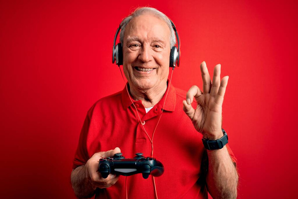 Senior grey haired gamer man playing video games using gamepad joystick over red background doing ok sign with fingers, excellent symbol - Foto, afbeelding