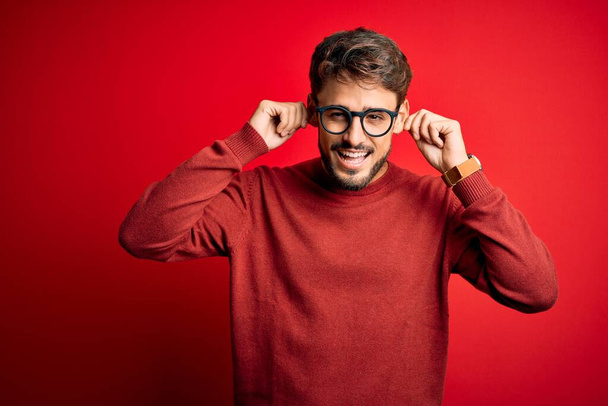 Młody przystojny mężczyzna w brodzie w okularach i swetrze stojący nad czerwonym tłem Uśmiechnięty ciągnący uszy palcami, zabawny gest. Problem przesłuchań - Zdjęcie, obraz