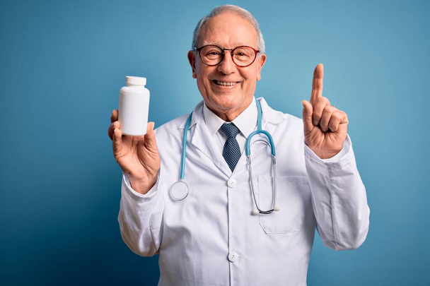 Hombre médico de pelo gris mayor sosteniendo píldoras farmacéuticas sobre fondo azul sorprendido con una idea o pregunta apuntando con el dedo con la cara feliz, número uno
 - Foto, imagen