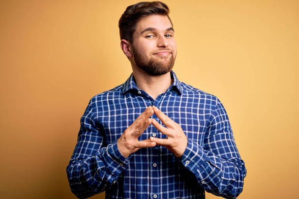Young blond businessman with beard and blue eyes wearing shirt over yellow background Hands together and fingers crossed smiling relaxed and cheerful. Success and optimistic - Foto, afbeelding