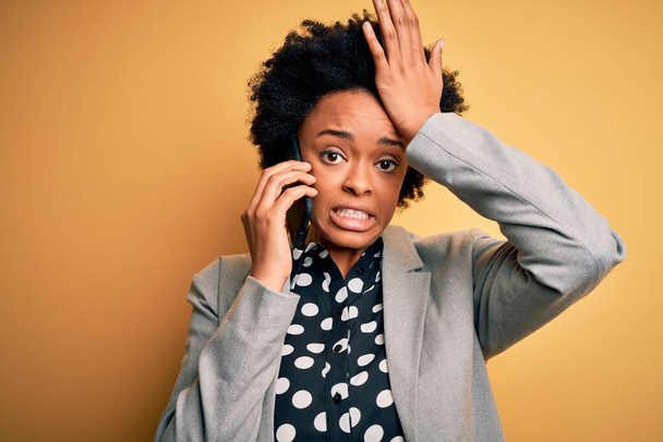 Young African American afro woman with curly hair having conversation talking on smartphone stressed with hand on head, shocked with shame and surprise face, angry and frustrated. Fear and upset for mistake. - 写真・画像