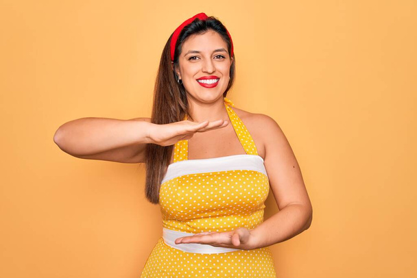 Young hispanic pin up woman wearing fashion sexy 50s style over yellow background gesturing with hands showing big and large size sign, measure symbol. Smiling looking at the camera. Measuring concept. - Foto, imagen