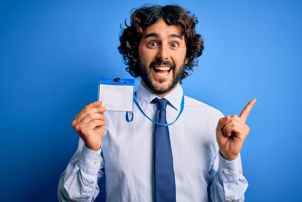 Jonge knappe zakenman met baard met ID-kaart identificatie over blauwe achtergrond zeer gelukkig wijzend met hand en vinger naar de zijkant - Foto, afbeelding